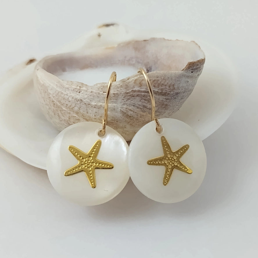 Gold starfish earrings on white stones with a shell in the background