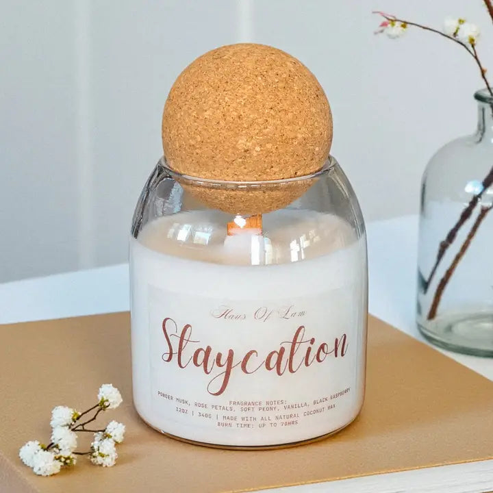 Candle in a glass jar with a cork lid on a wooden surface, surrounded by small white flowers.