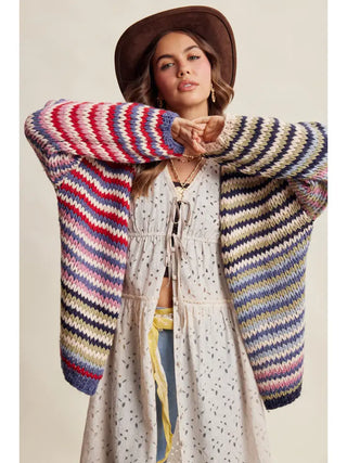 A woman wearing a chunky knit bohemian cardigan with multicolor stripes in red, blue, yellow, green, and cream, styled over a white boho dress and a brown wide-brim hat against a solid beige background.
