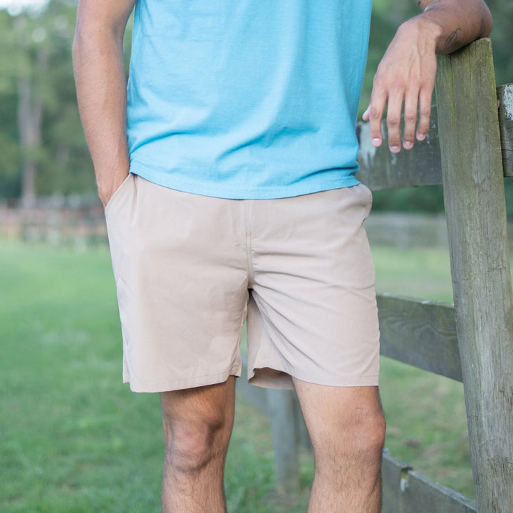 Person wearing a blue shirt and beige shorts standing by a wooden fence with a grassy area and trees in the background.