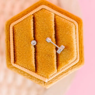 A close-up of a silver post and matching stud earring displayed on a textured mustard-yellow jewelry box.