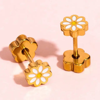 A pair of gold flower-shaped stud earrings with white enamel petals and a small yellow center, displayed on a soft pink background.