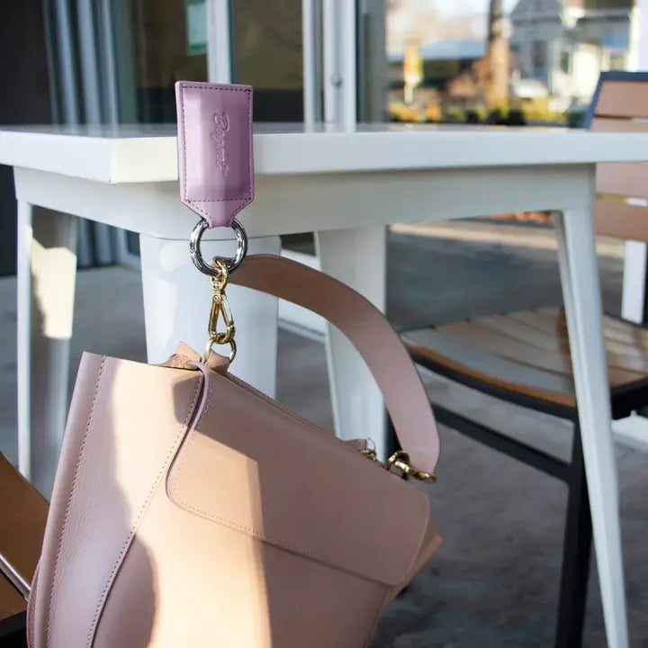 Pink handbag with a purple strap and keychain attached, set against an outdoor cafe background.