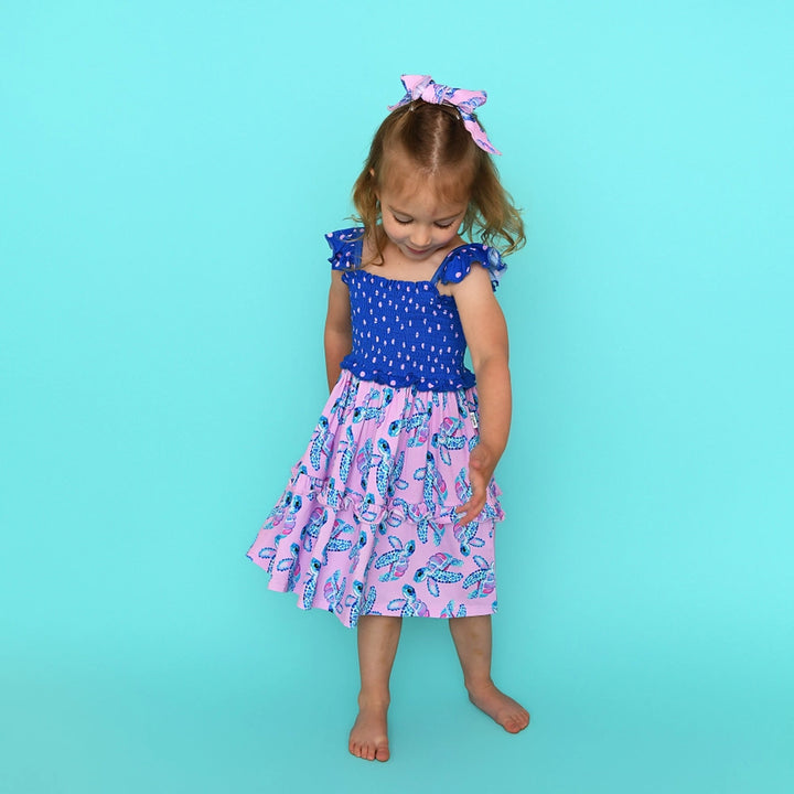 Young girl wearing a blue top and pink dress with turtle pattern on a teal background