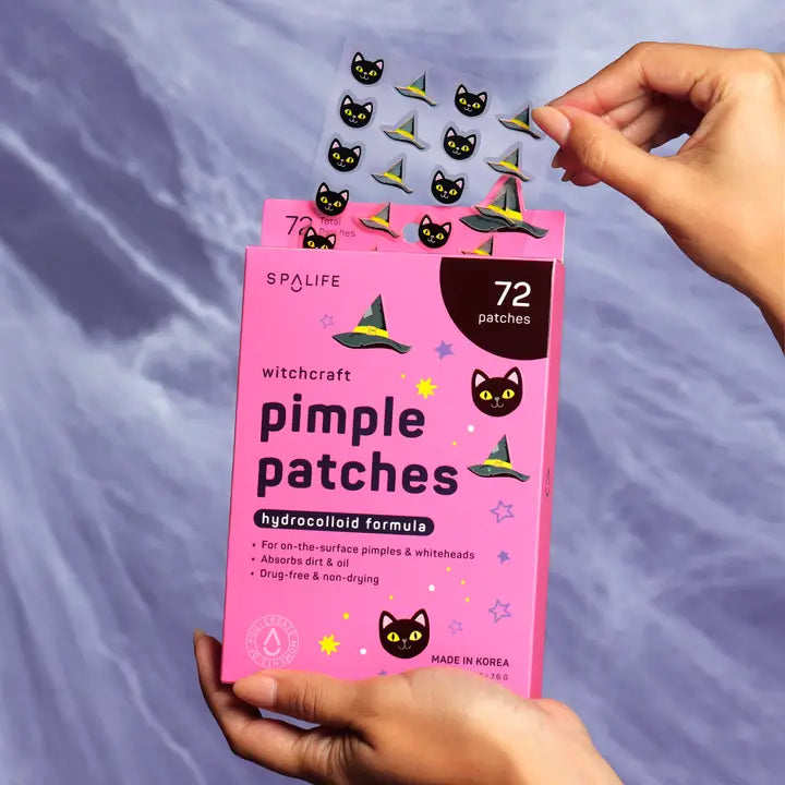 Pink packaging of pimple patches with black cat and witch hat designs, held by a hand against a purple fabric background.