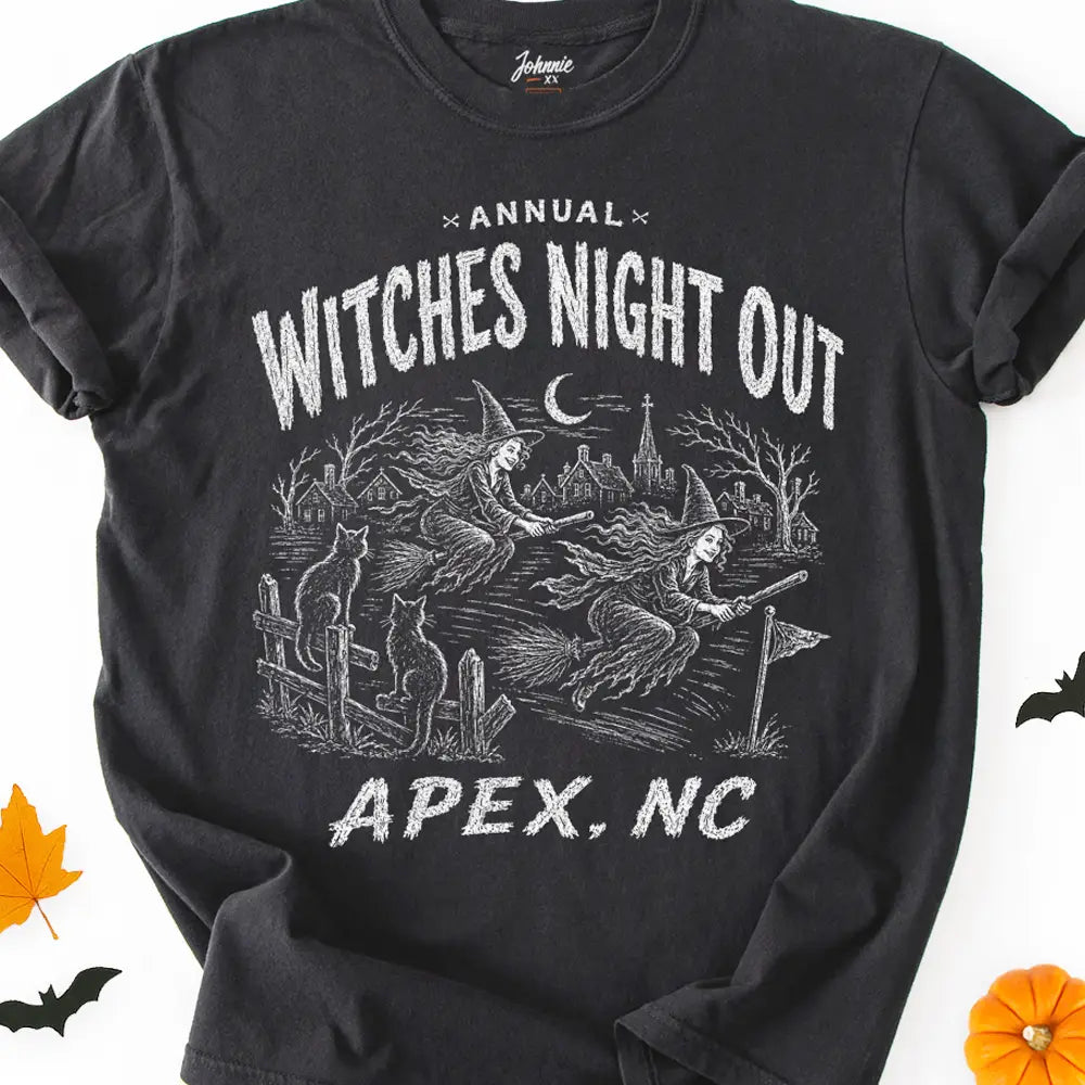 A black graphic t-shirt laid flat on a white background with Halloween-themed props like a pumpkin, maple leaf, and bats. The shirt features a white distressed graphic that reads 'Annual Witches Night Out APEX, NC' with an illustration of witches riding broomsticks over a graveyard.