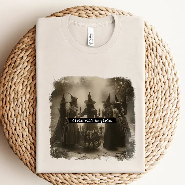 A folded off-white t-shirt with a vintage-style black and white graphic of five girls in witch hats, displayed on a round woven straw mat.