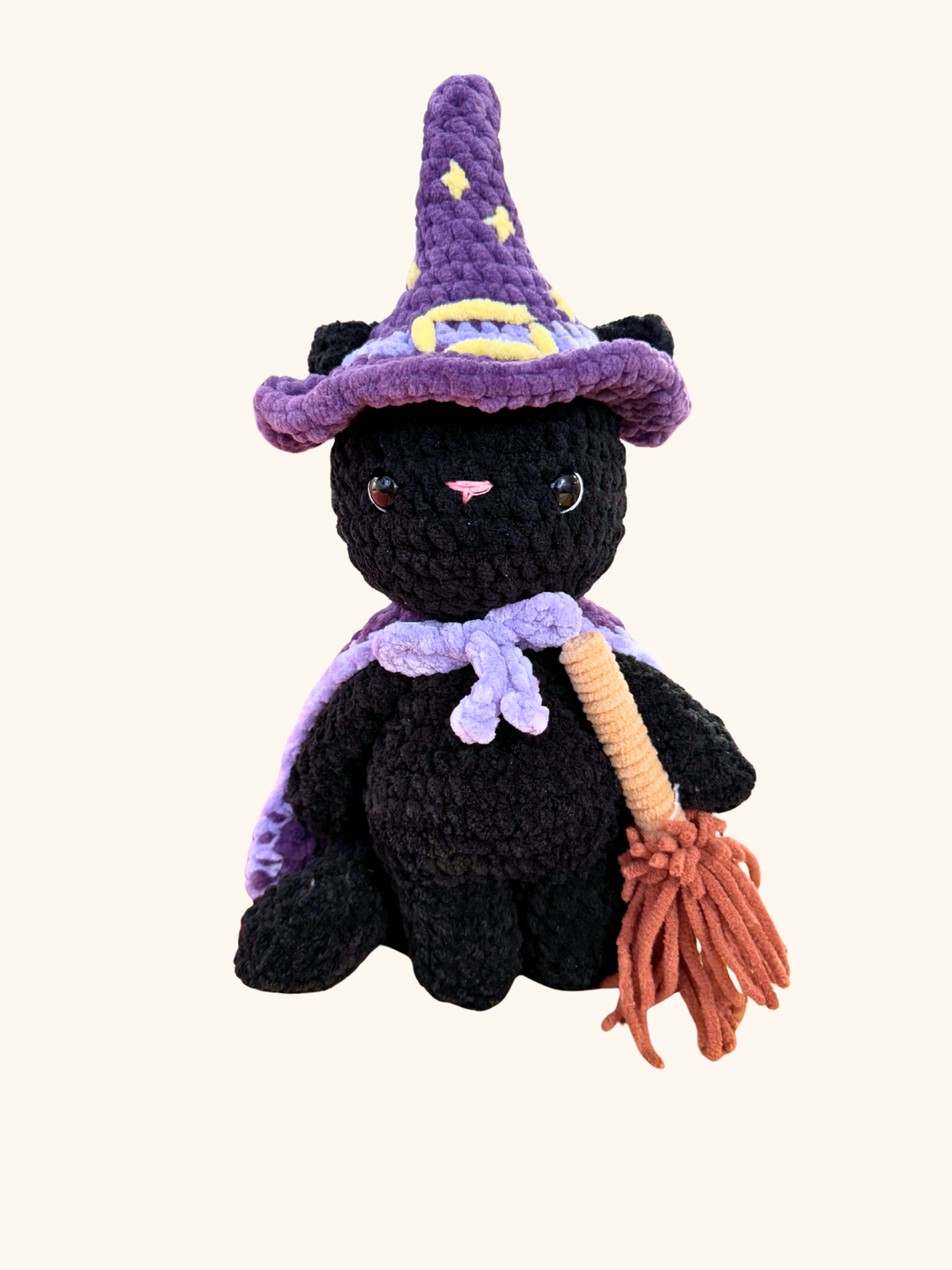 Crocheted black cat toy with a purple witch hat and broomstick on a white background