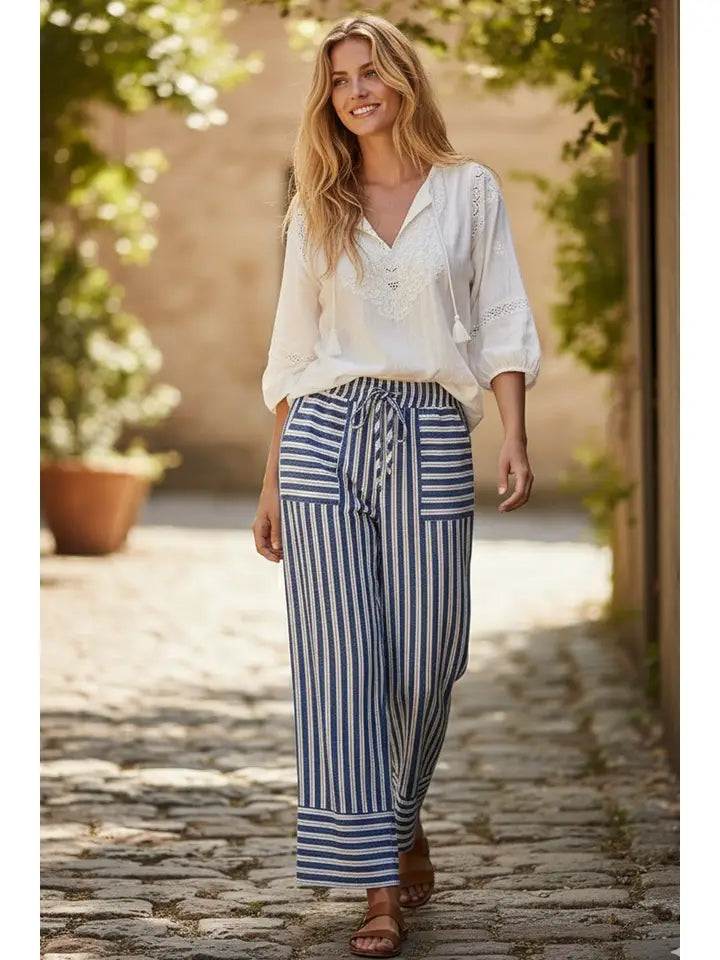 Woman wearing a white blouse and blue striped pants standing on a stone path with greenery.
