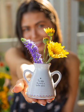 Person holding a small white vase with yellow and purple flowers outdoors.
