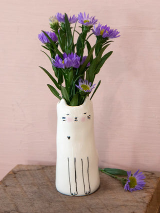 Cat-shaped vase with purple flowers on a wooden surface against a pink background