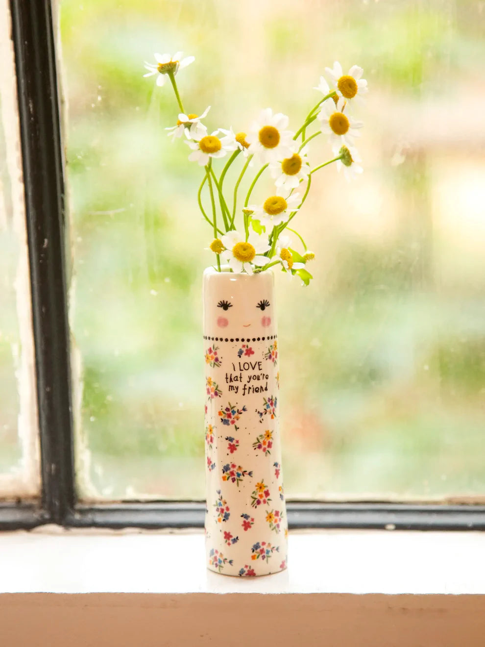 Vase with floral design and face on a windowsill with flowers.