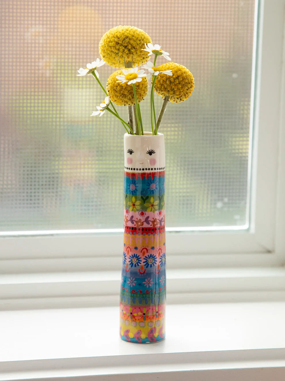 Colorful vase with a face design holding flowers on a windowsill.