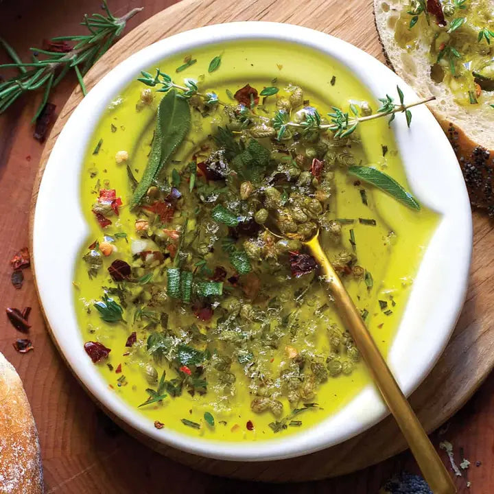 A bowl of herb-flavored dip mix, surrounded by bread, herbs, and a drizzle of olive oil.