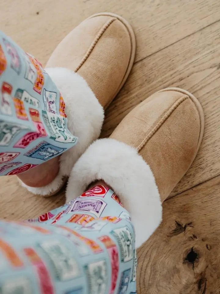 Tan slippers with white fur lining worn by a person in colorful pajama pants on a wooden floor.